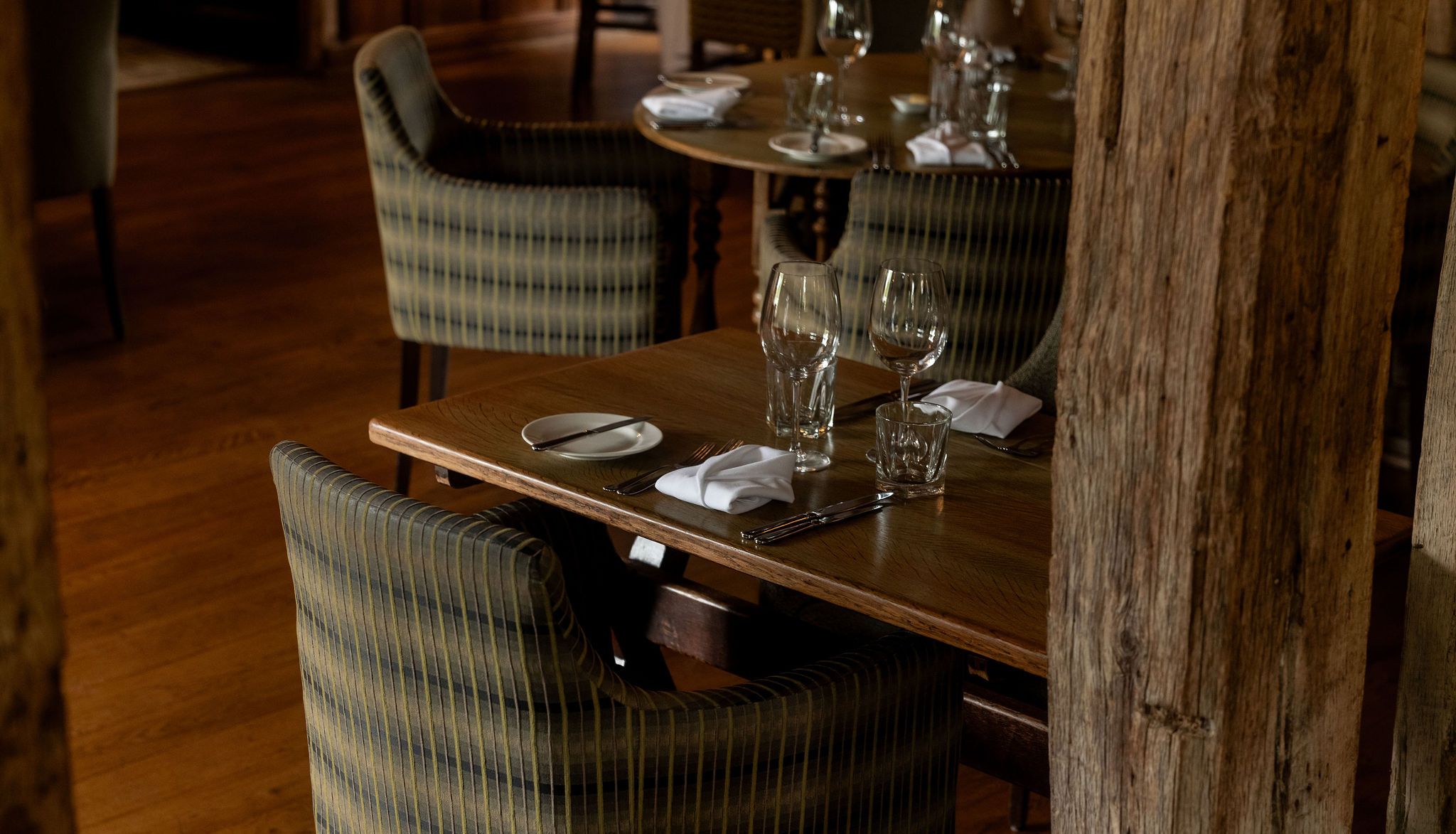 A cozy restaurant setting with wooden tables set for two, featuring striped cushioned chairs, glassware, napkins, and cutlery, on a wood floor with rustic wooden beams in the foreground.