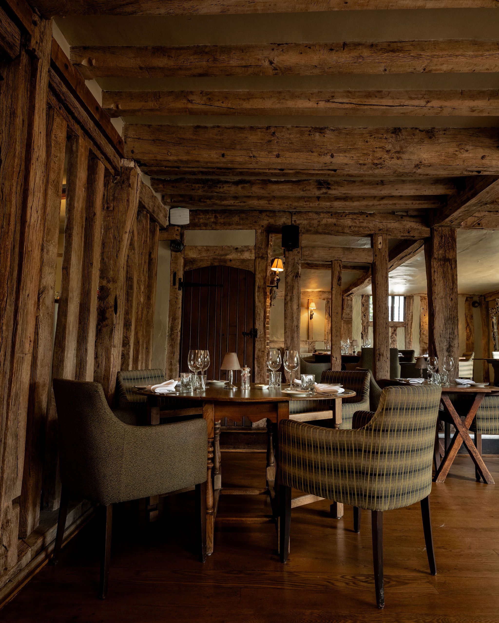 Rustic restaurant interior with wooden beams, cozy upholstered chairs, and neatly set tables with glassware and napkins.