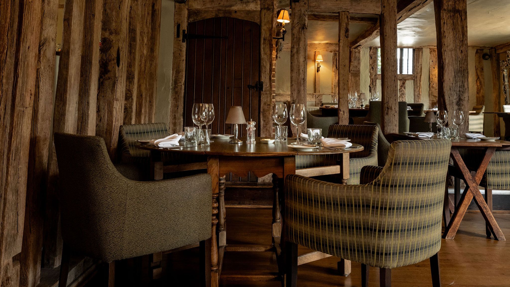Rustic restaurant interior with wooden beams, cozy upholstered chairs, and neatly set tables with glassware and napkins.