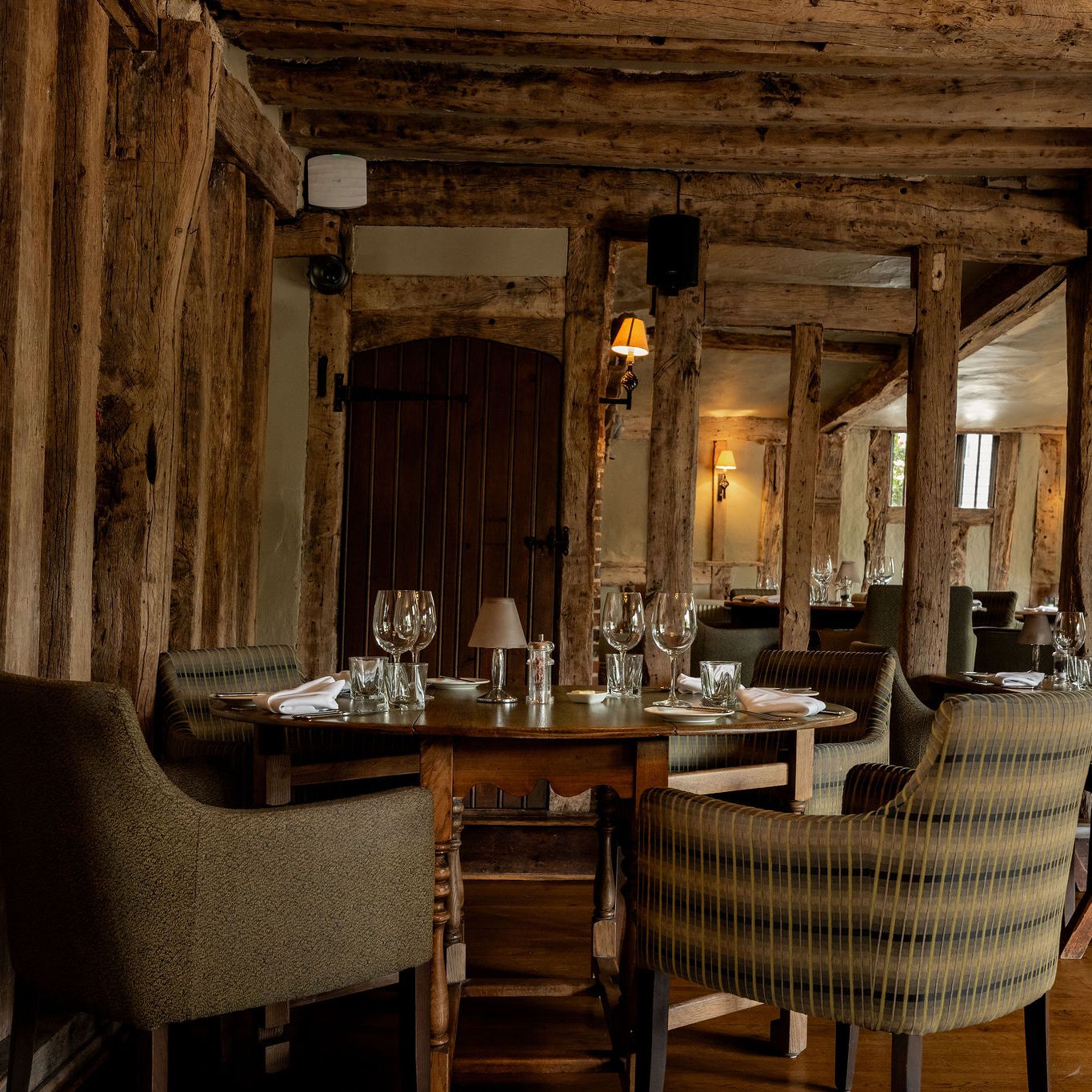 Rustic restaurant interior with wooden beams, round tables set with glassware and napkins, and upholstered chairs.