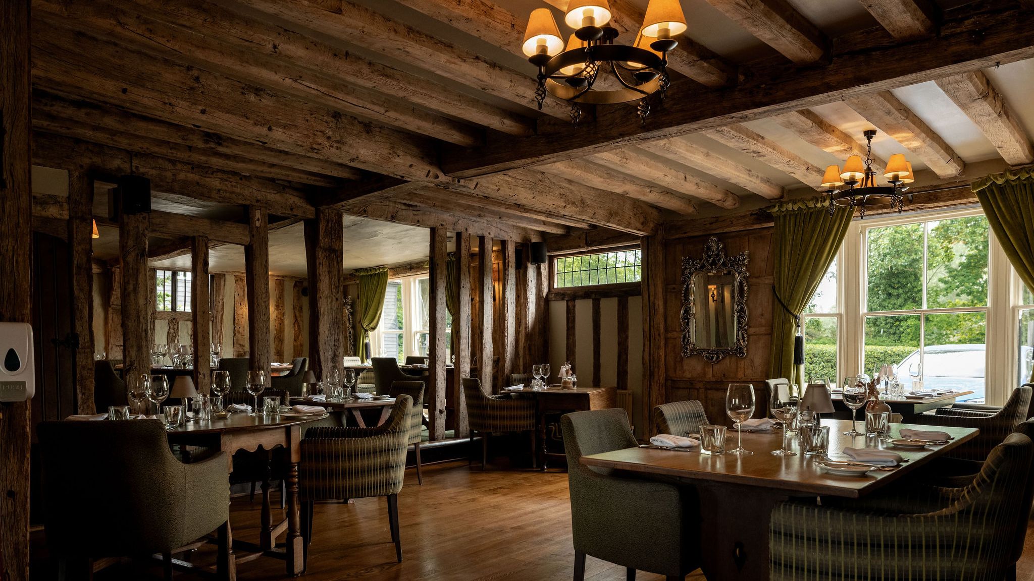 Cozy restaurant interior with rustic wooden beams, elegant lighting, green curtains, and tables set with glassware and napkins near large windows.