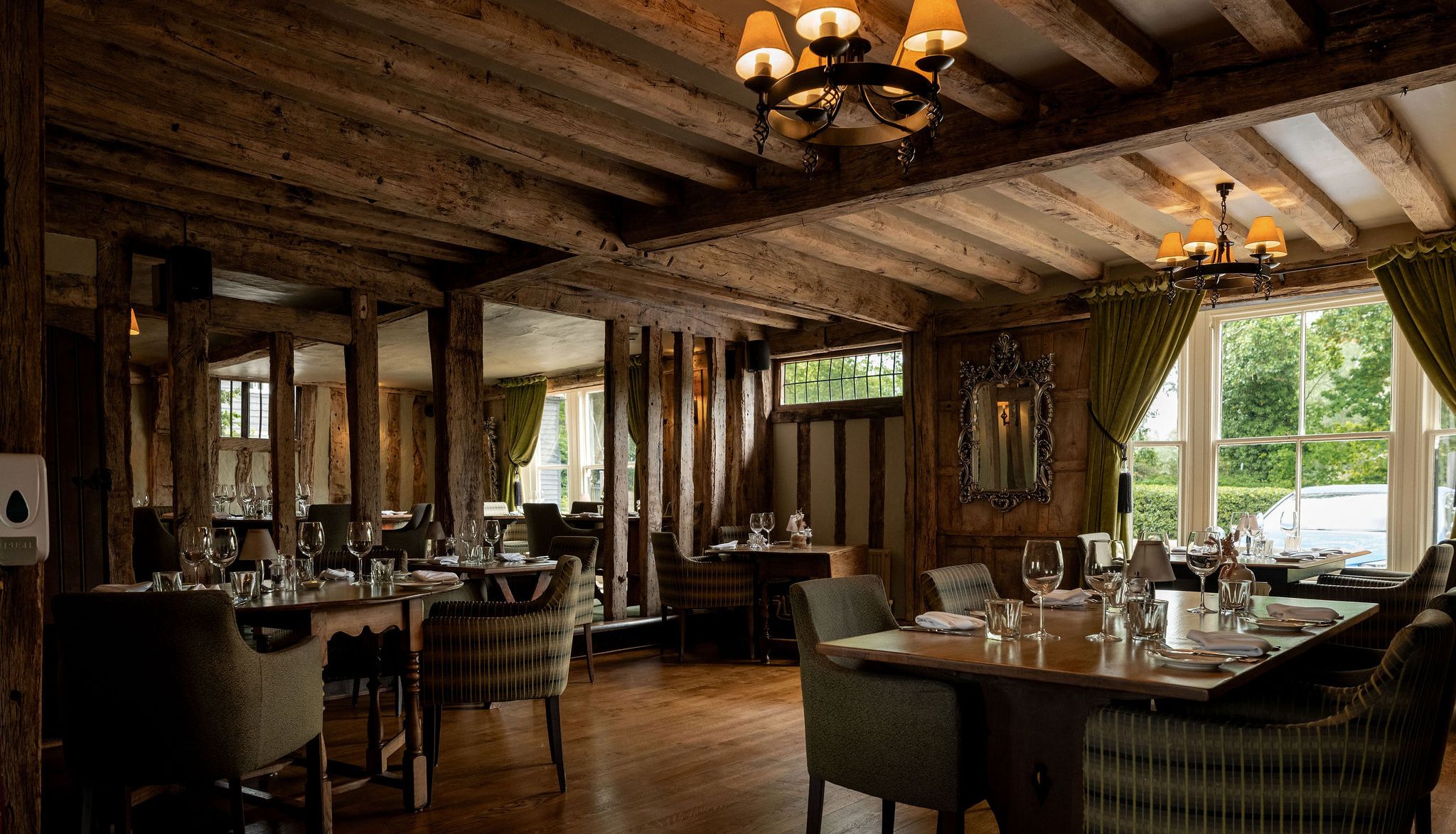 Cozy restaurant interior with rustic wooden beams, elegant lighting, green curtains, and tables set with glassware and napkins near large windows.