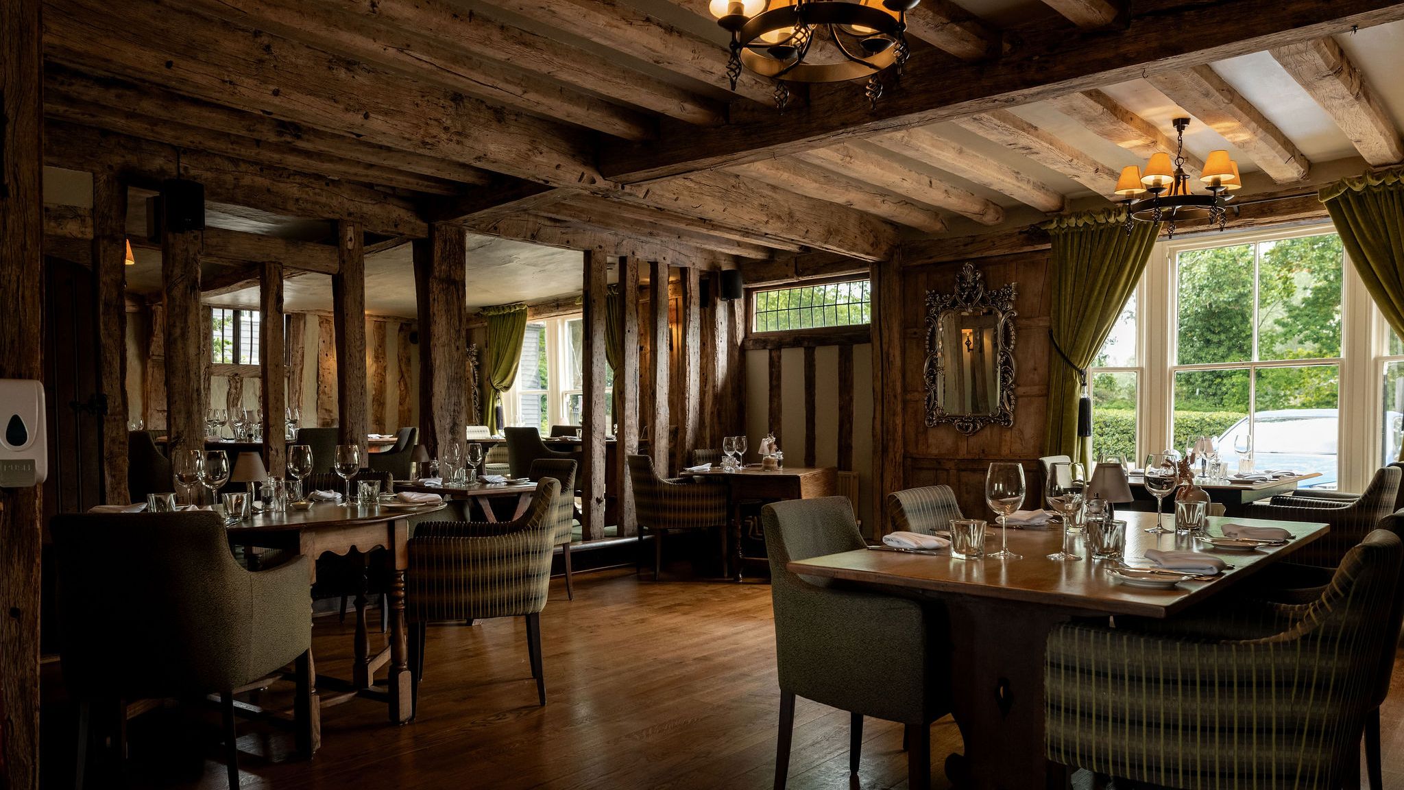 Cozy restaurant interior with rustic wooden beams, elegant lighting, green curtains, and tables set with glassware and napkins near large windows.