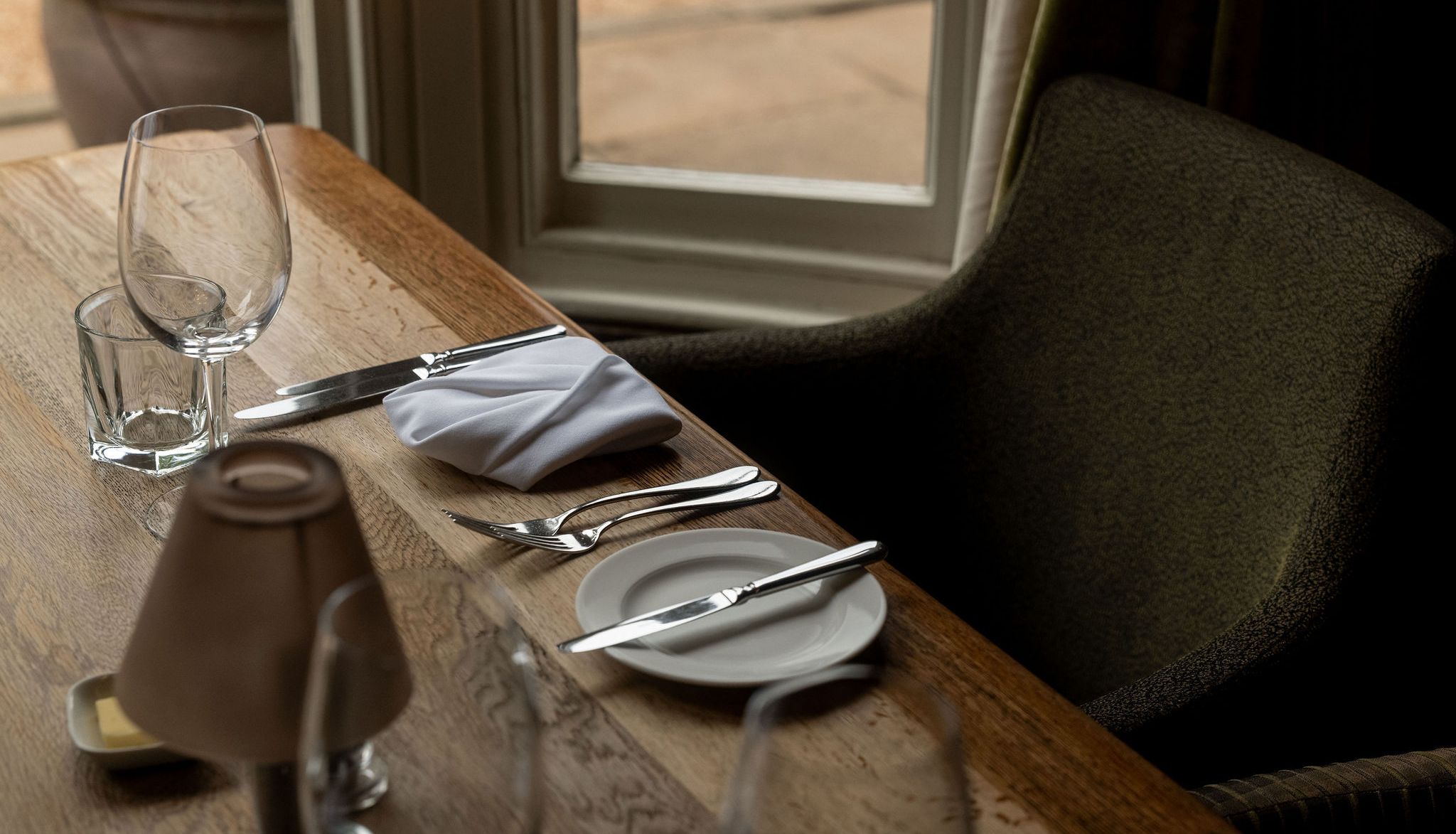 A close-up of a neatly set dining table by a window, featuring a wine glass, water glass, silverware, a plate with a butter knife, a folded white napkin, and a small lamp.