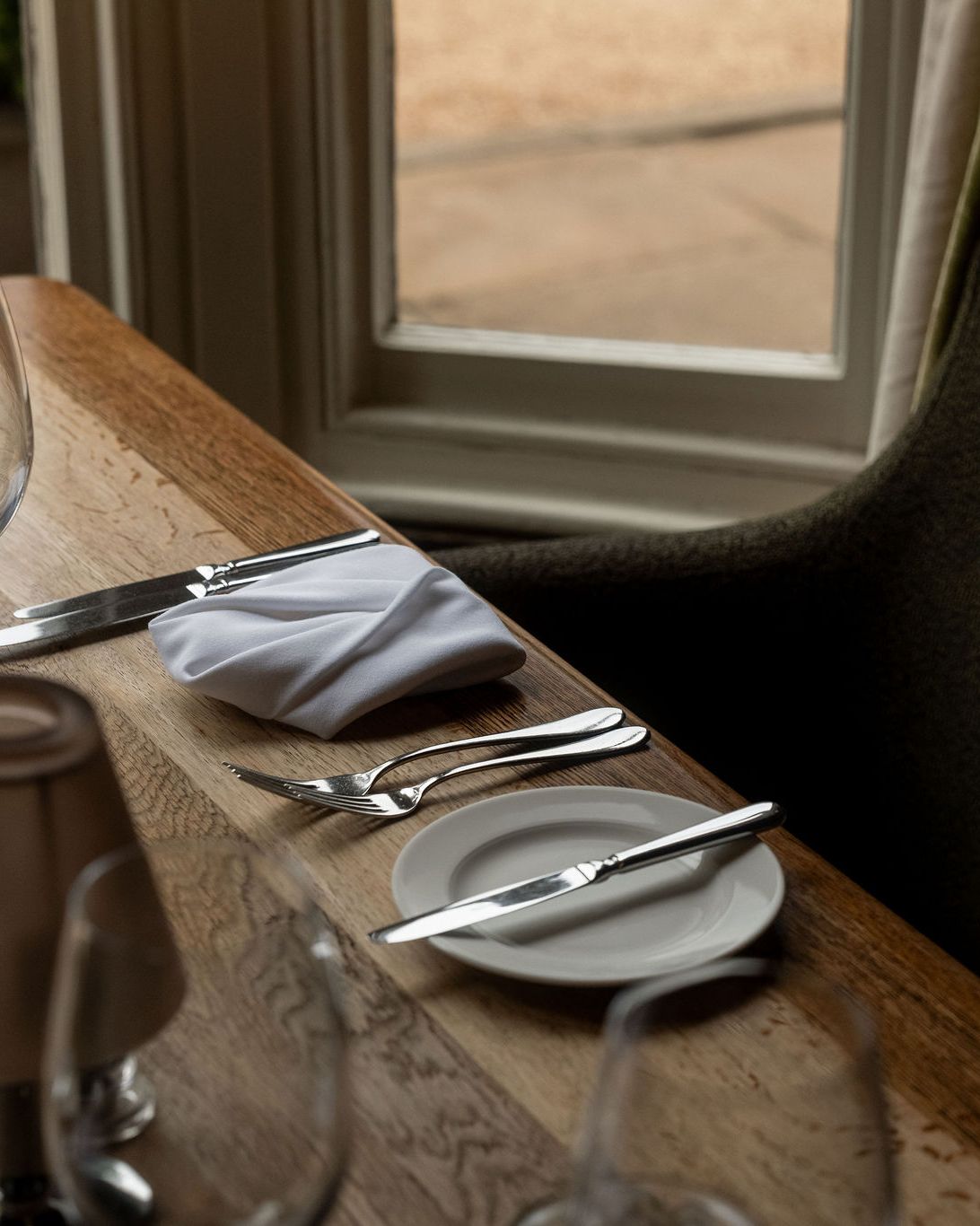 A close-up of a neatly set dining table by a window, featuring a wine glass, water glass, silverware, a plate with a butter knife, a folded white napkin, and a small lamp.