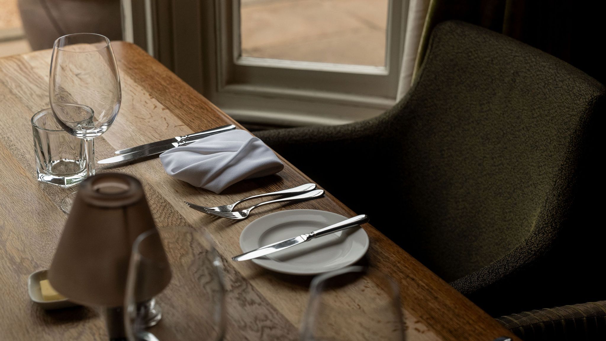 A close-up of a neatly set dining table by a window, featuring a wine glass, water glass, silverware, a plate with a butter knife, a folded white napkin, and a small lamp.
