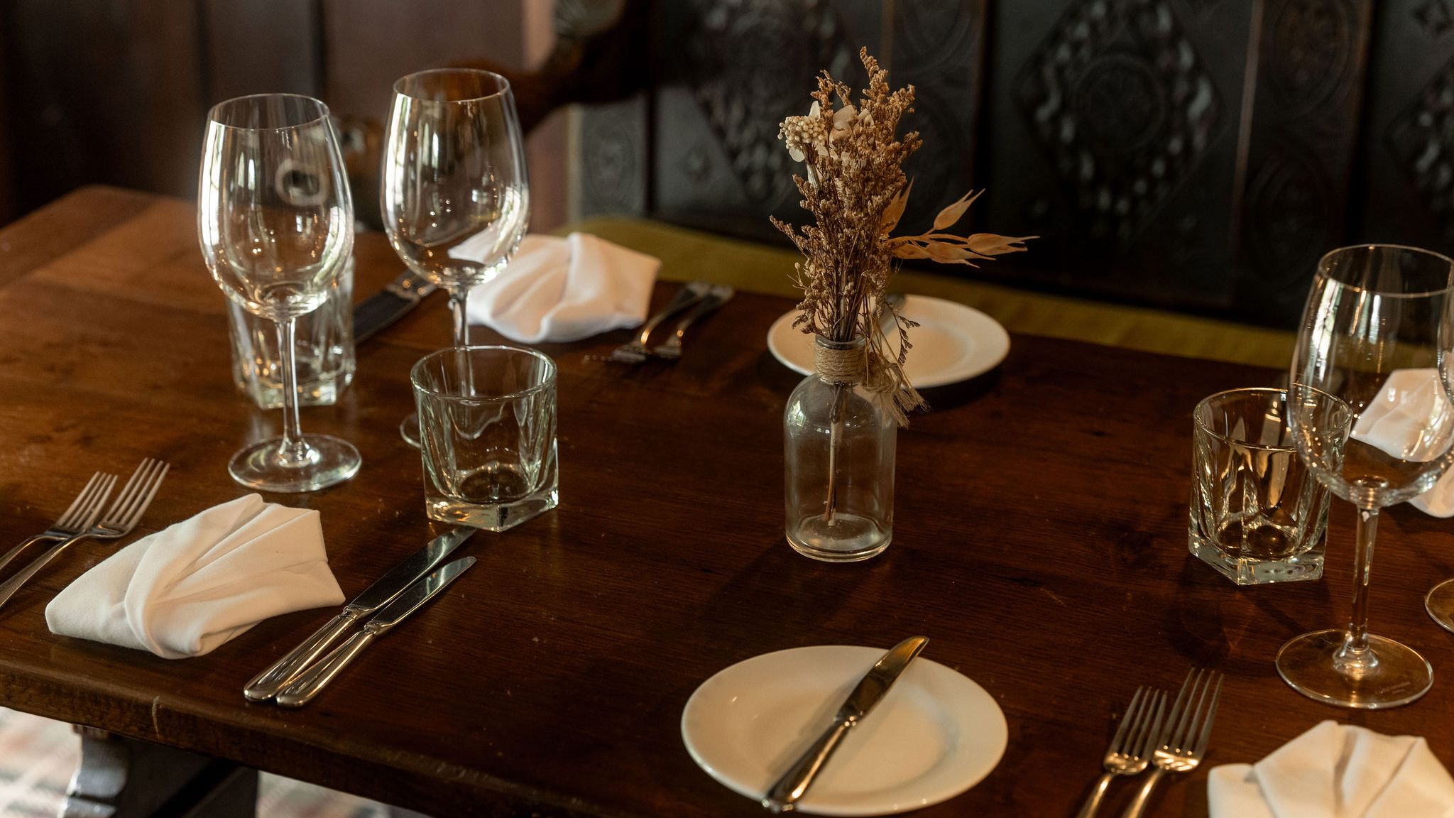 A rustic wooden dining table set for four with glassware, plates, silverware, white napkins, and a small vase containing dried flowers as a centerpiece.