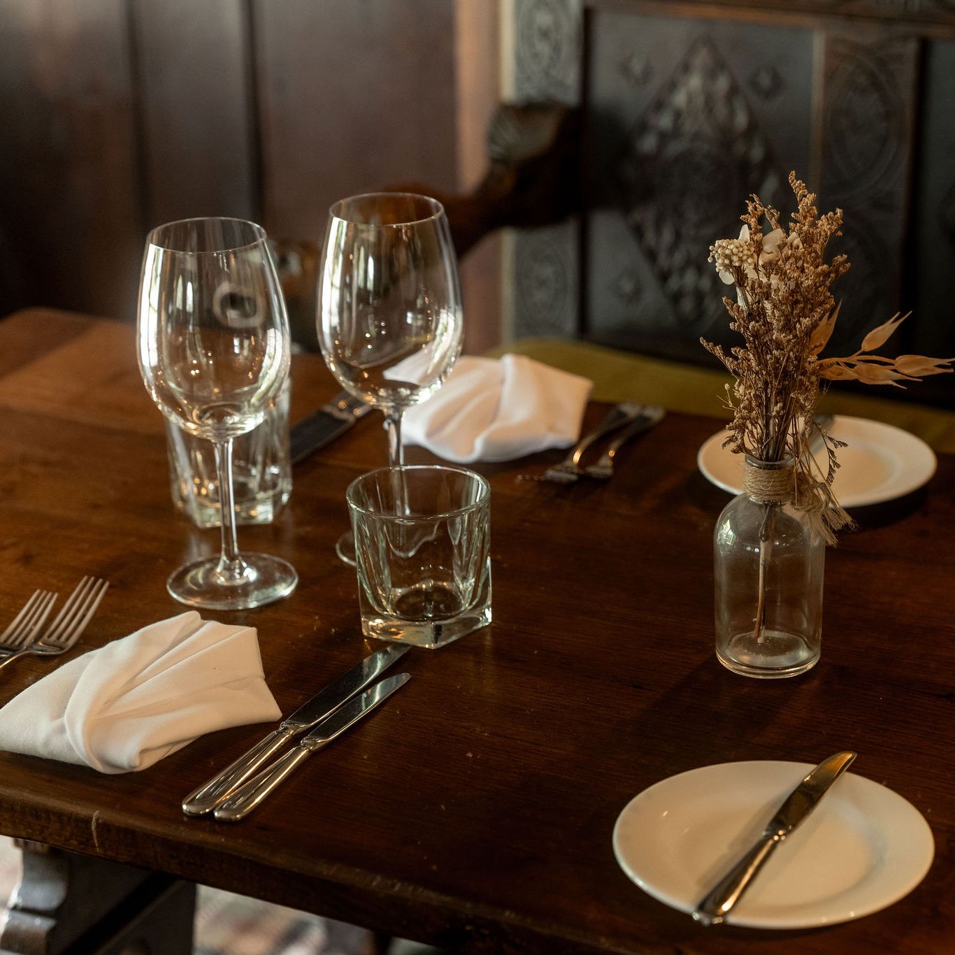 A rustic wooden dining table set for four with glassware, plates, silverware, white napkins, and a small vase containing dried flowers as a centerpiece.