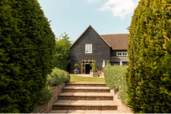 A modern barn-style house with dark wooden exterior, surrounded by manicured hedges and greenery, viewed from a brick pathway with steps leading up to the entrance.