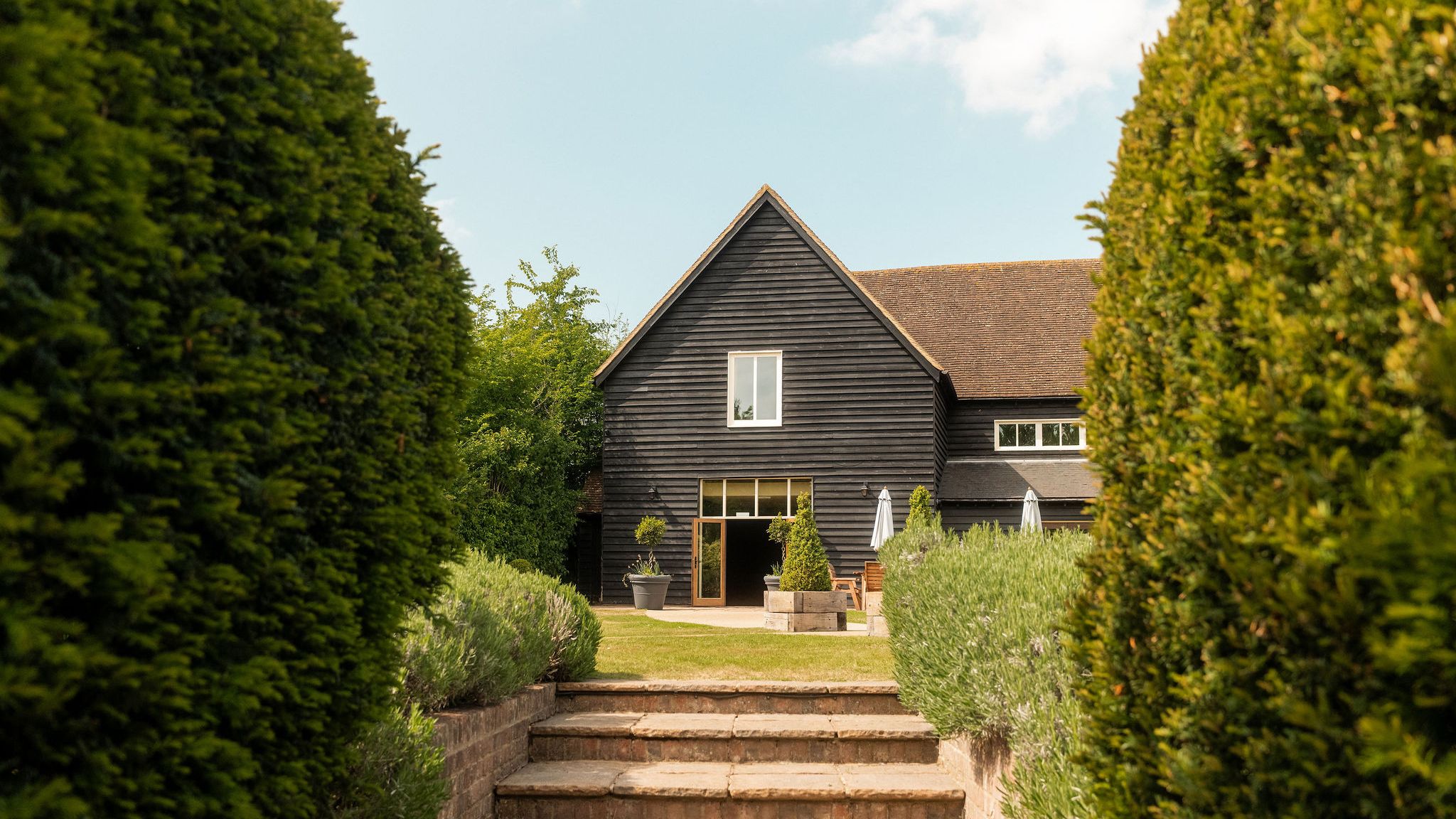 A modern barn-style house with dark wooden exterior, surrounded by manicured hedges and greenery, viewed from a brick pathway with steps leading up to the entrance.