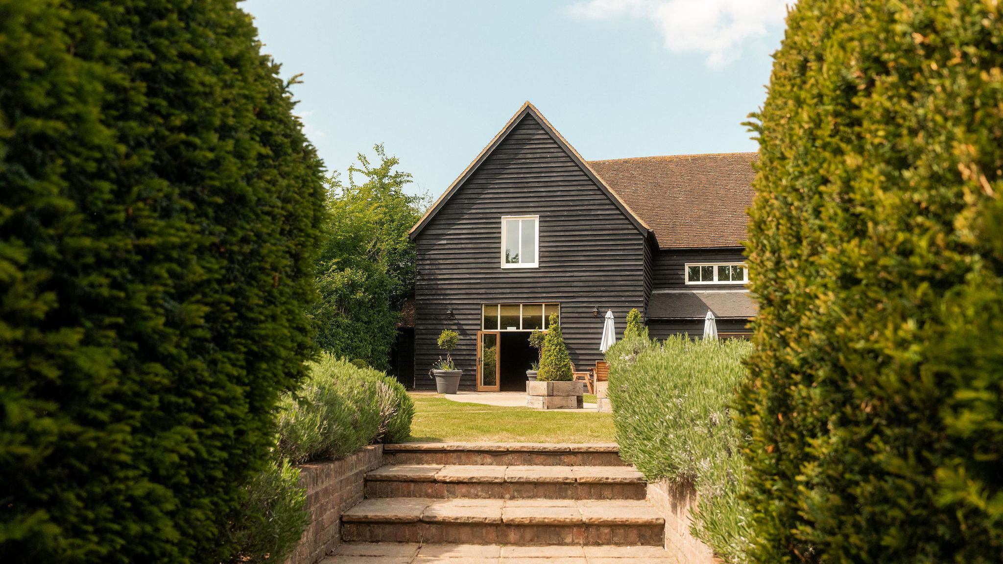 A modern barn-style house with dark wooden exterior, surrounded by manicured hedges and greenery, viewed from a brick pathway with steps leading up to the entrance.