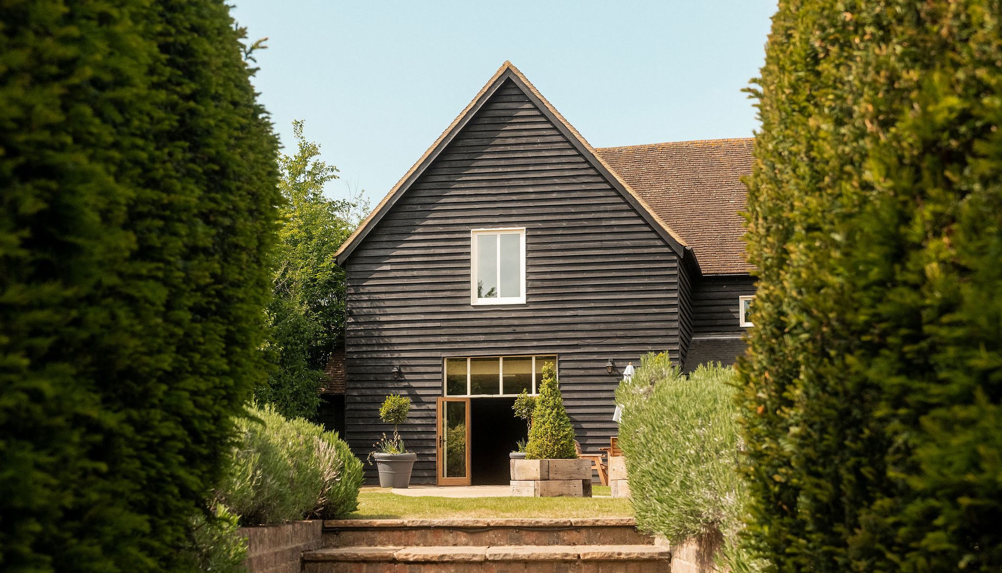 Stone steps leading up through manicured bushes to the entrance of a black wooden house with a gabled roof on a sunny day.