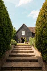 Stone steps leading up through manicured bushes to the entrance of a black wooden house with a gabled roof on a sunny day.