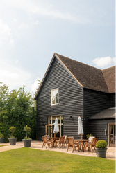 Modern black wooden house with sloped roof, outdoor patio with wooden tables, chairs, umbrellas, and potted plants on a sunny day.