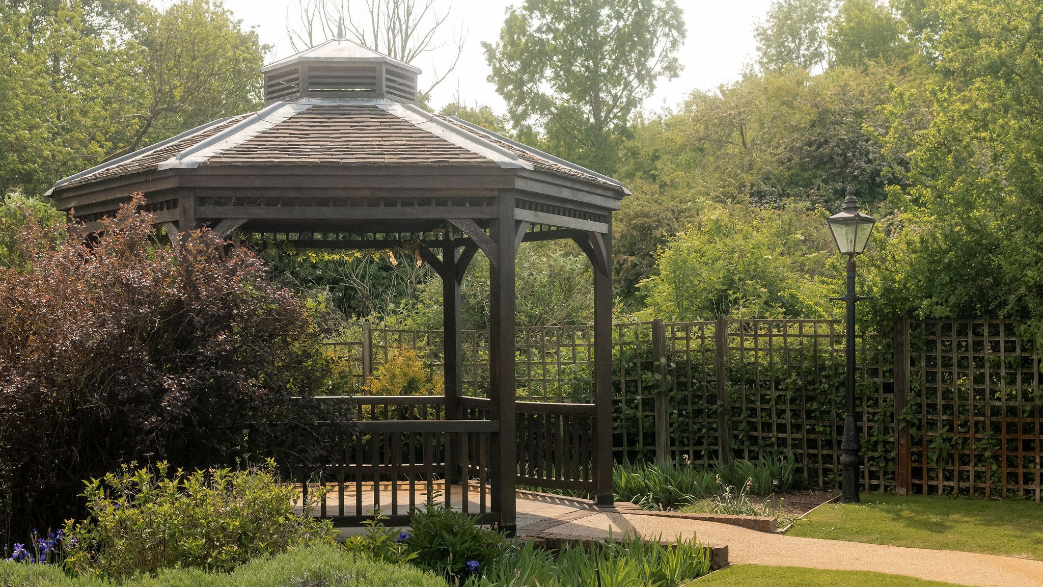 Wooden gazebo in a landscaped garden surrounded by green bushes, flowers, and trees with a pathway and a lamppost nearby.