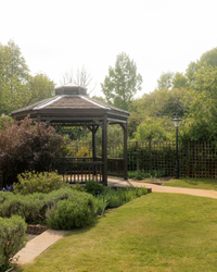 Wooden gazebo in a landscaped garden surrounded by green bushes, flowers, and trees with a pathway and a lamppost nearby.