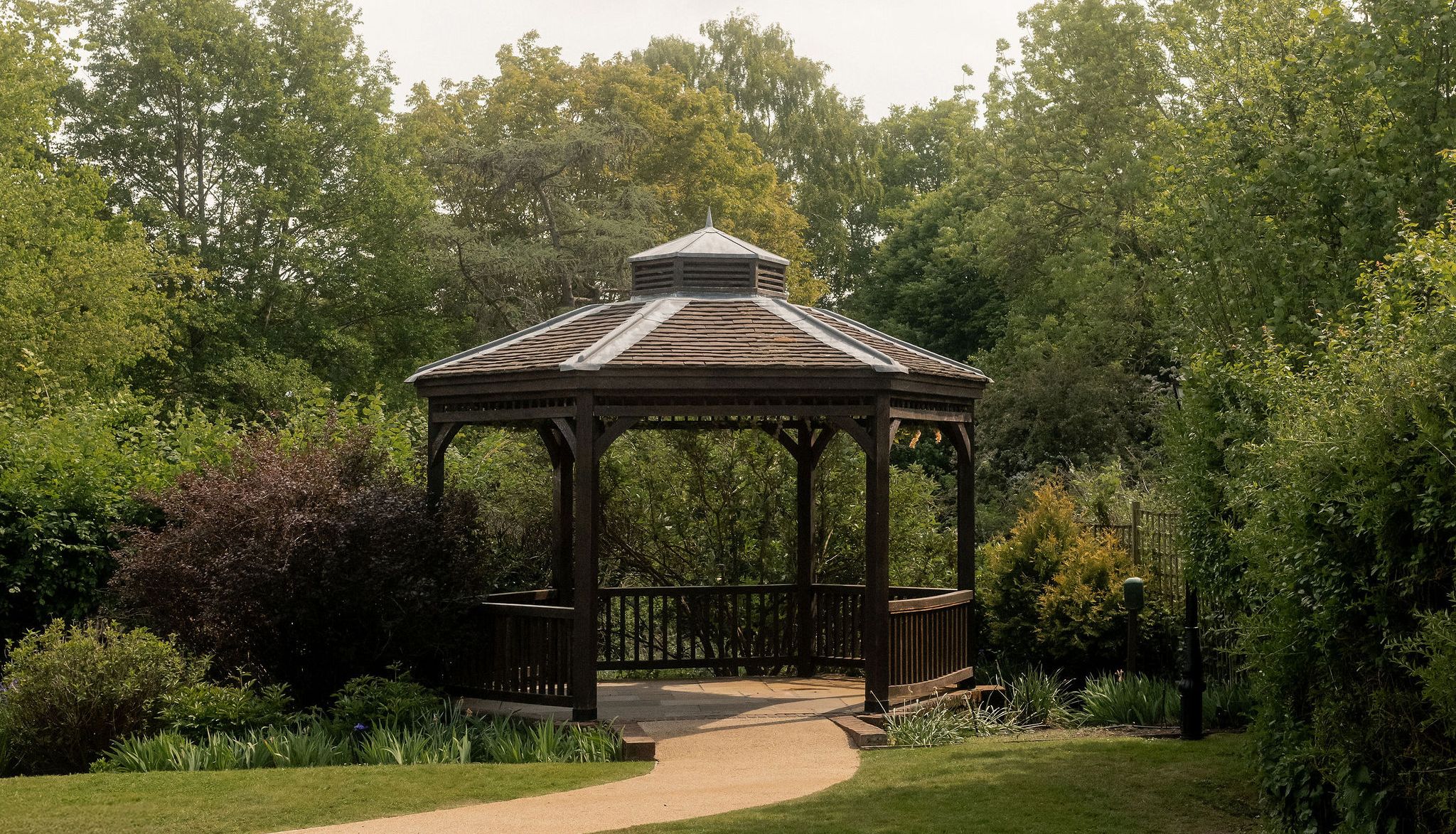 A wooden gazebo surrounded by greenery and trees, with a curved path leading to it in a garden setting.