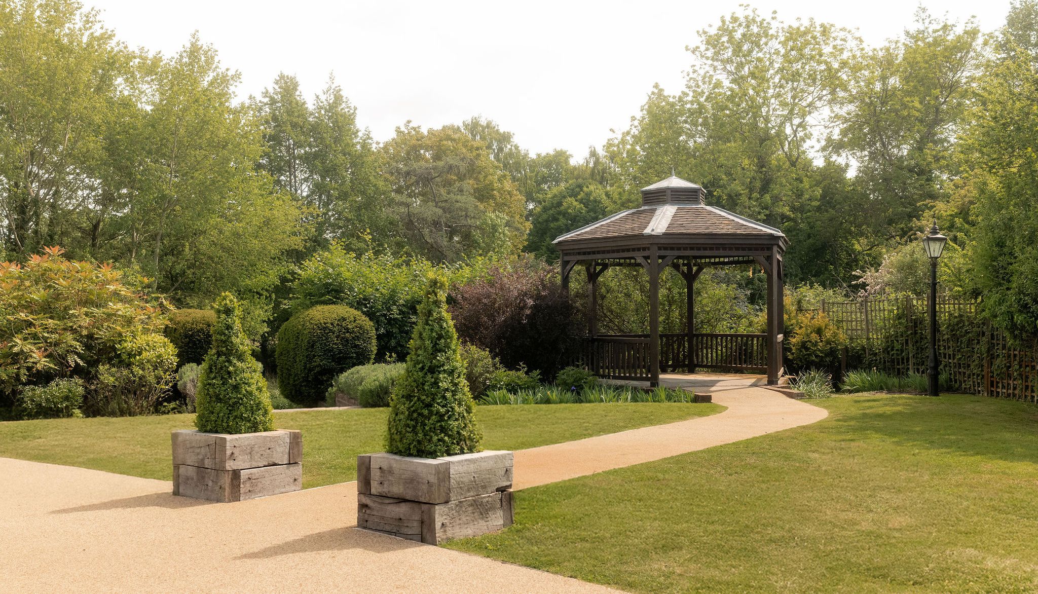 A peaceful garden with a wooden gazebo, neatly trimmed hedges, green trees, a paved pathway, and decorative planter boxes with conical shrubs.
