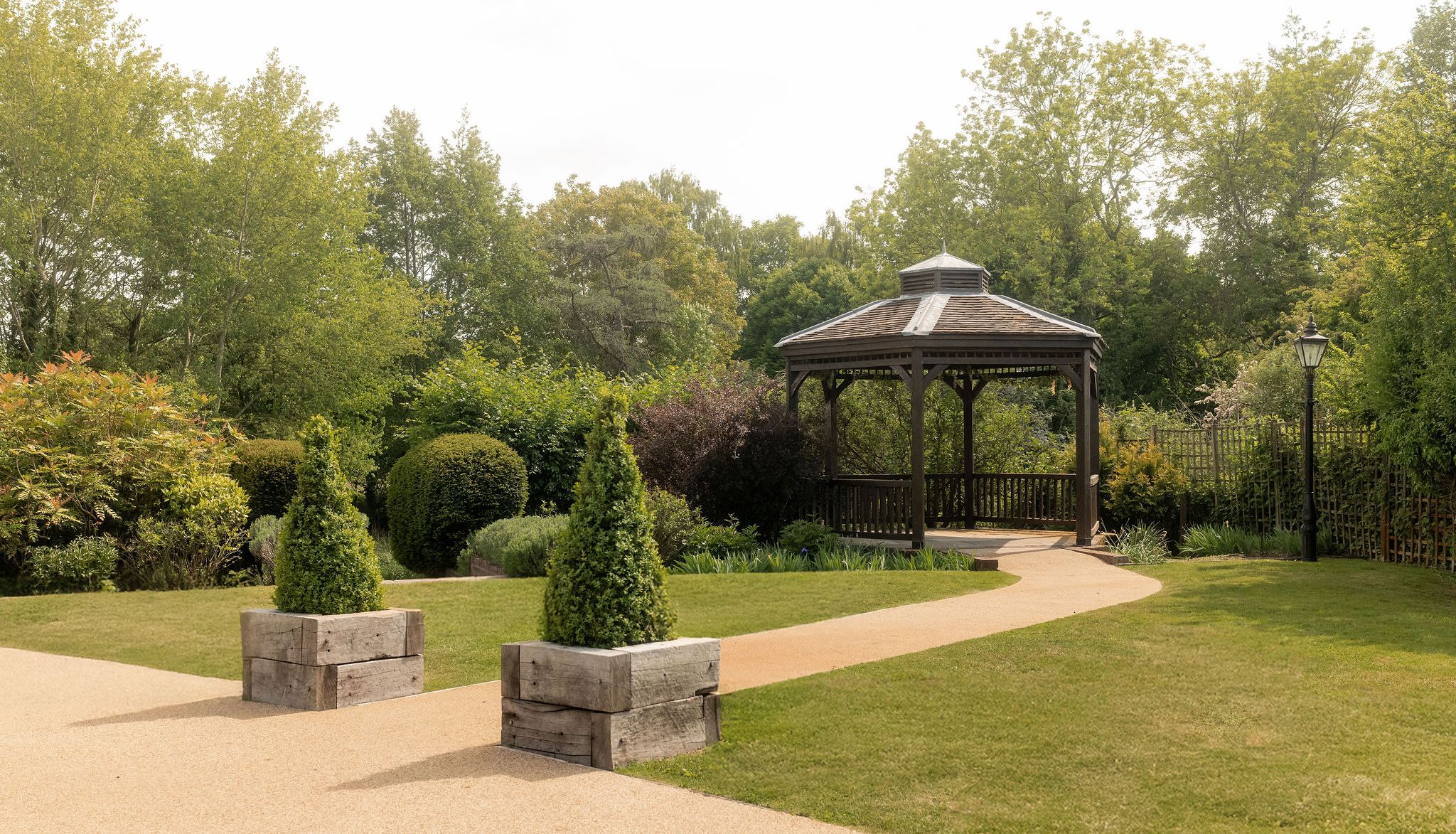 A peaceful garden with a wooden gazebo, neatly trimmed hedges, green trees, a paved pathway, and decorative planter boxes with conical shrubs.