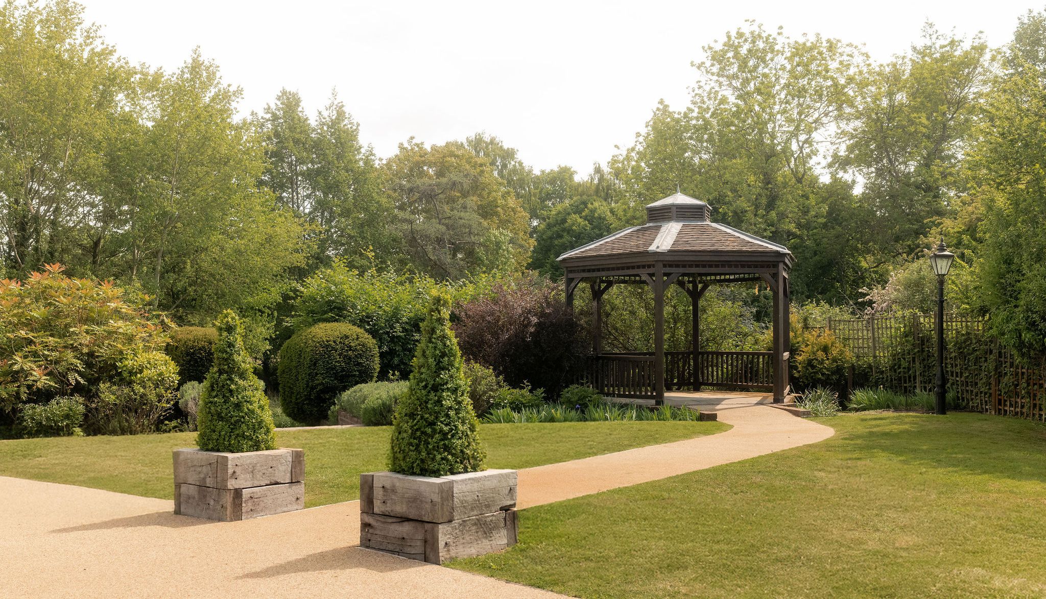 A peaceful garden with a wooden gazebo, neatly trimmed hedges, green trees, a paved pathway, and decorative planter boxes with conical shrubs.