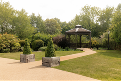 A peaceful garden with a wooden gazebo, neatly trimmed hedges, green trees, a paved pathway, and decorative planter boxes with conical shrubs.