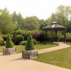 A peaceful garden with a wooden gazebo, neatly trimmed hedges, green trees, a paved pathway, and decorative planter boxes with conical shrubs.