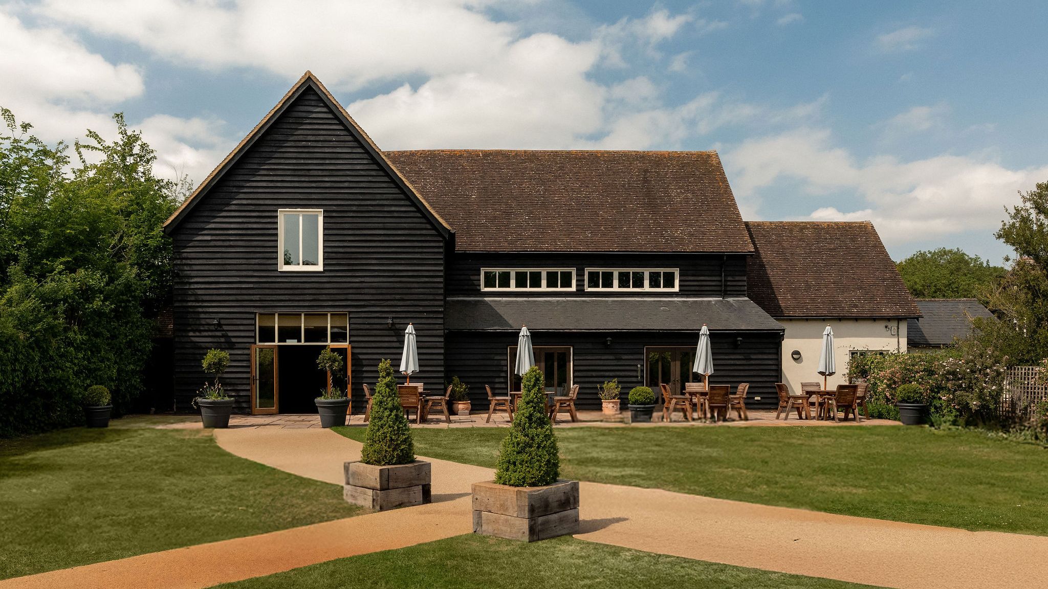 A large black wooden barn with a sloped roof, outdoor seating with tables and umbrellas, surrounded by well-kept grass, shrubbery, and a clear sky with scattered clouds.
