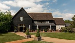 A large black wooden barn with a sloped roof, outdoor seating with tables and umbrellas, surrounded by well-kept grass, shrubbery, and a clear sky with scattered clouds.