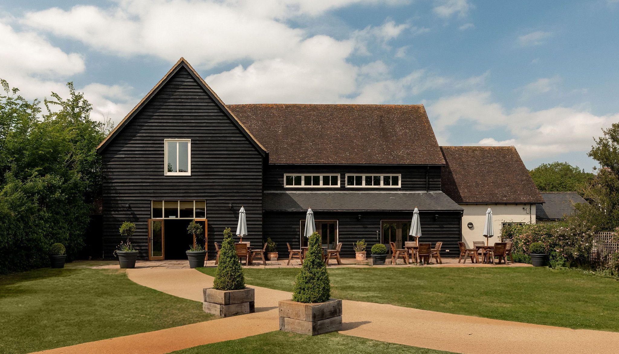 A large black wooden barn with a sloped roof, outdoor seating with tables and umbrellas, surrounded by well-kept grass, shrubbery, and a clear sky with scattered clouds.