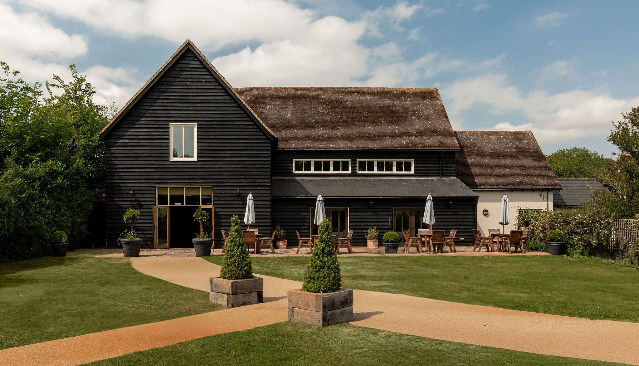 A large black wooden barn with a sloped roof, outdoor seating with tables and umbrellas, surrounded by well-kept grass, shrubbery, and a clear sky with scattered clouds.