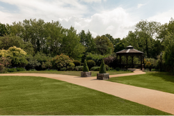 A landscaped garden with green grass, bushes, and trees. There is a paved pathway that leads to a wooden gazebo on the right side of the image.