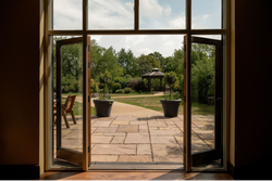 Open glass doors leading to a stone patio with two large potted plants and a view of a garden gazebo surrounded by greenery.