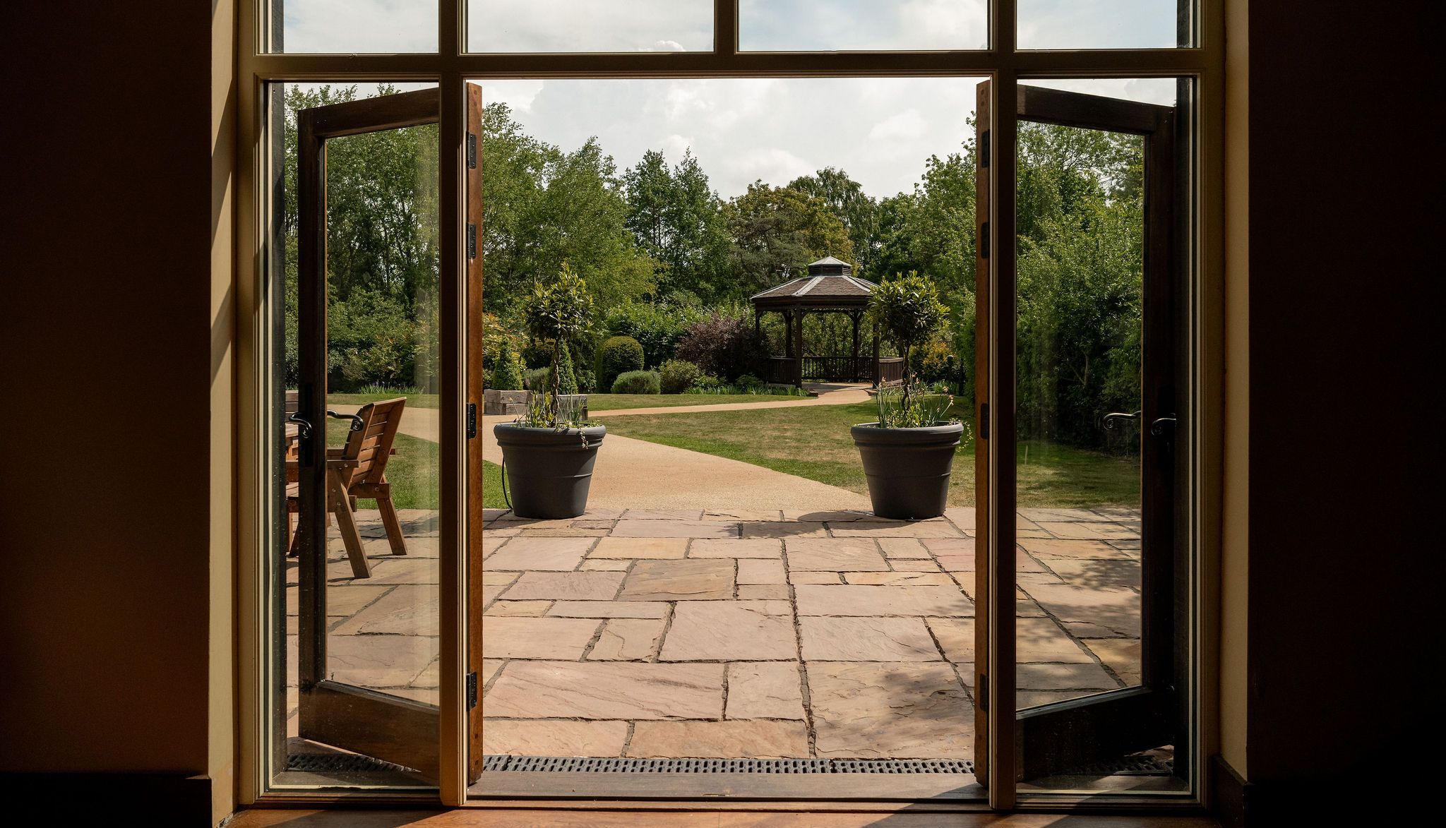 Open glass doors leading to a stone patio with two large potted plants and a view of a garden gazebo surrounded by greenery.