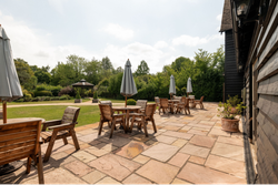 Outdoor patio area with wooden tables and chairs, large umbrellas, and a stone tile floor, overlooking a lush garden with green trees and a gazebo in the background.