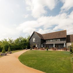 A modern barn-style building with outdoor seating, surrounded by green lawn and a curved pathway under a partly cloudy sky.