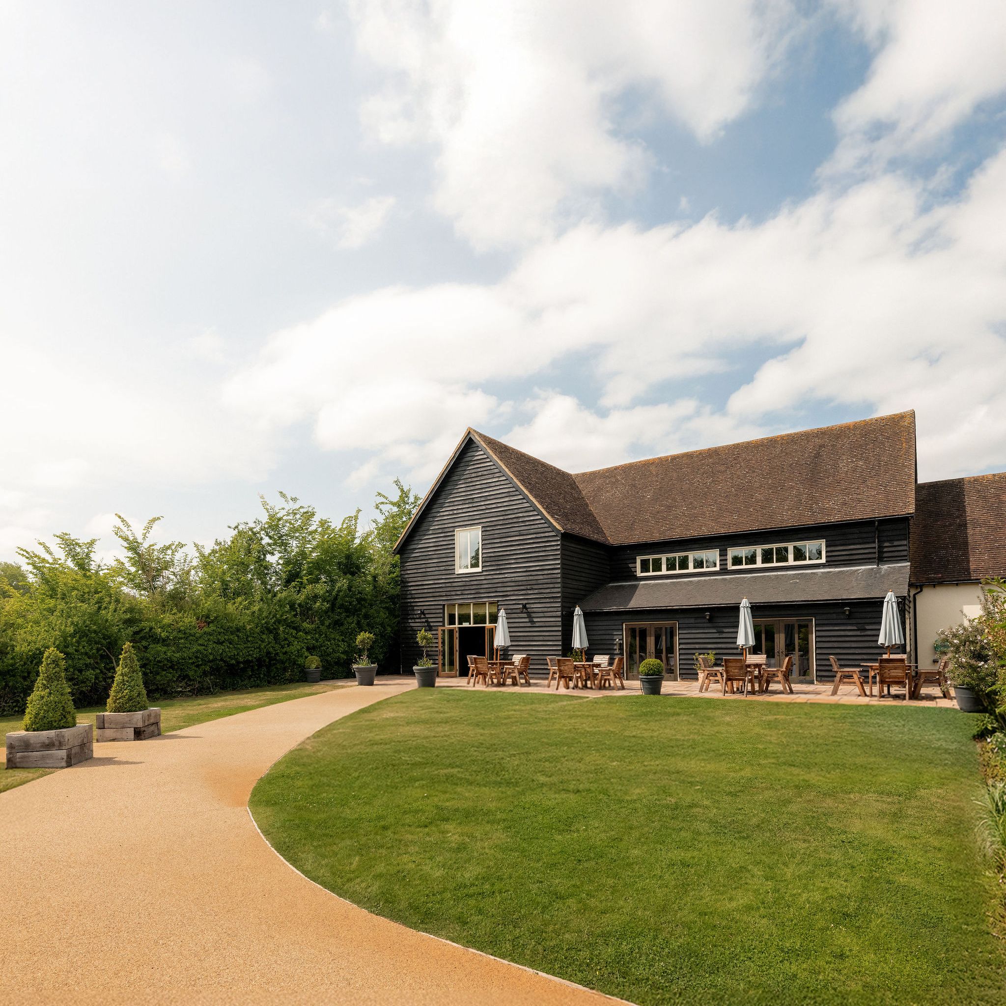 A modern barn-style building with outdoor seating, surrounded by green lawn and a curved pathway under a partly cloudy sky.