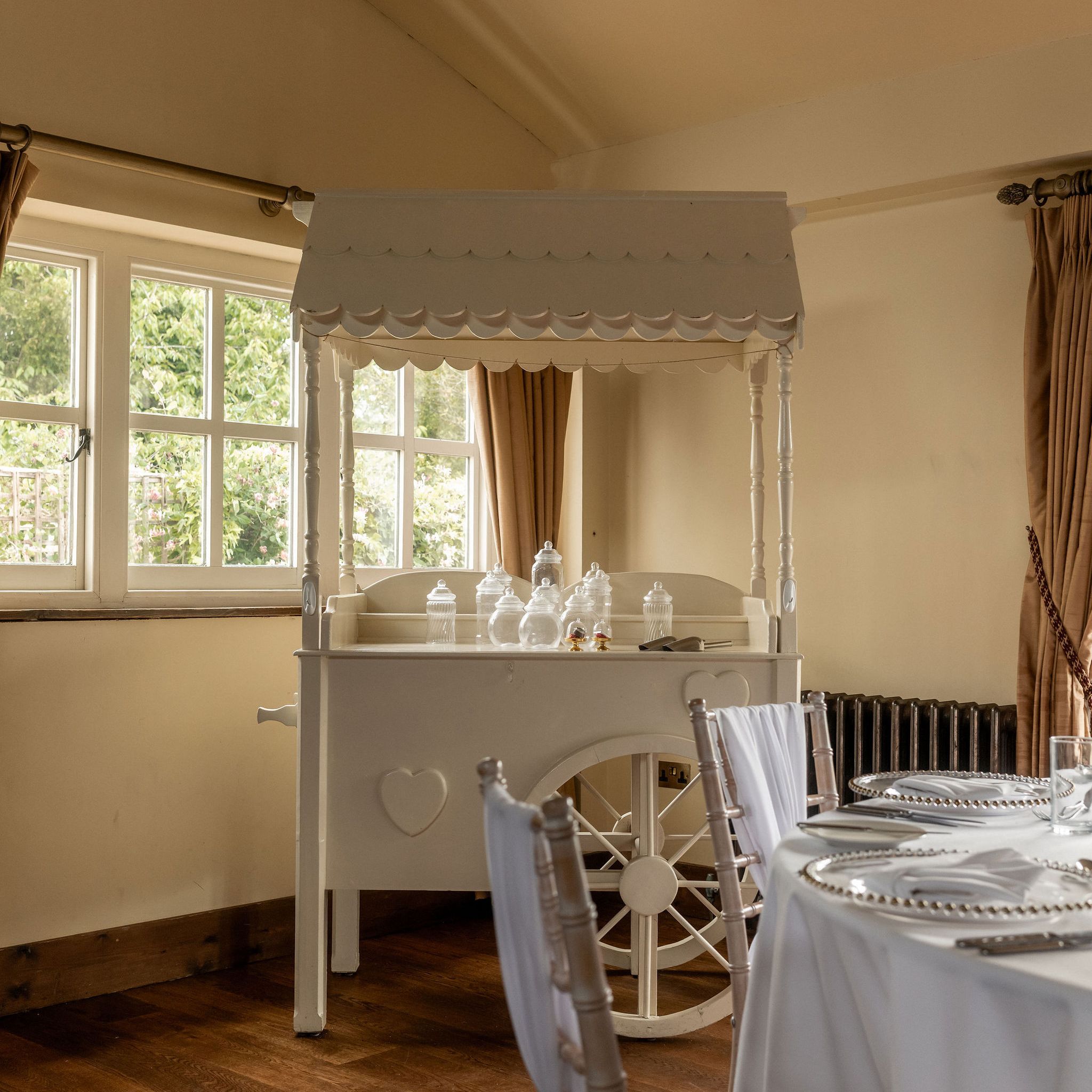 Elegant indoor setting with a vintage white candy cart, decorated with glass jars, and a dining table set with white tablecloth, napkins, and silver plates near a window with brown curtains.