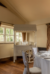 Elegant indoor setting with a vintage white candy cart, decorated with glass jars, and a dining table set with white tablecloth, napkins, and silver plates near a window with brown curtains.