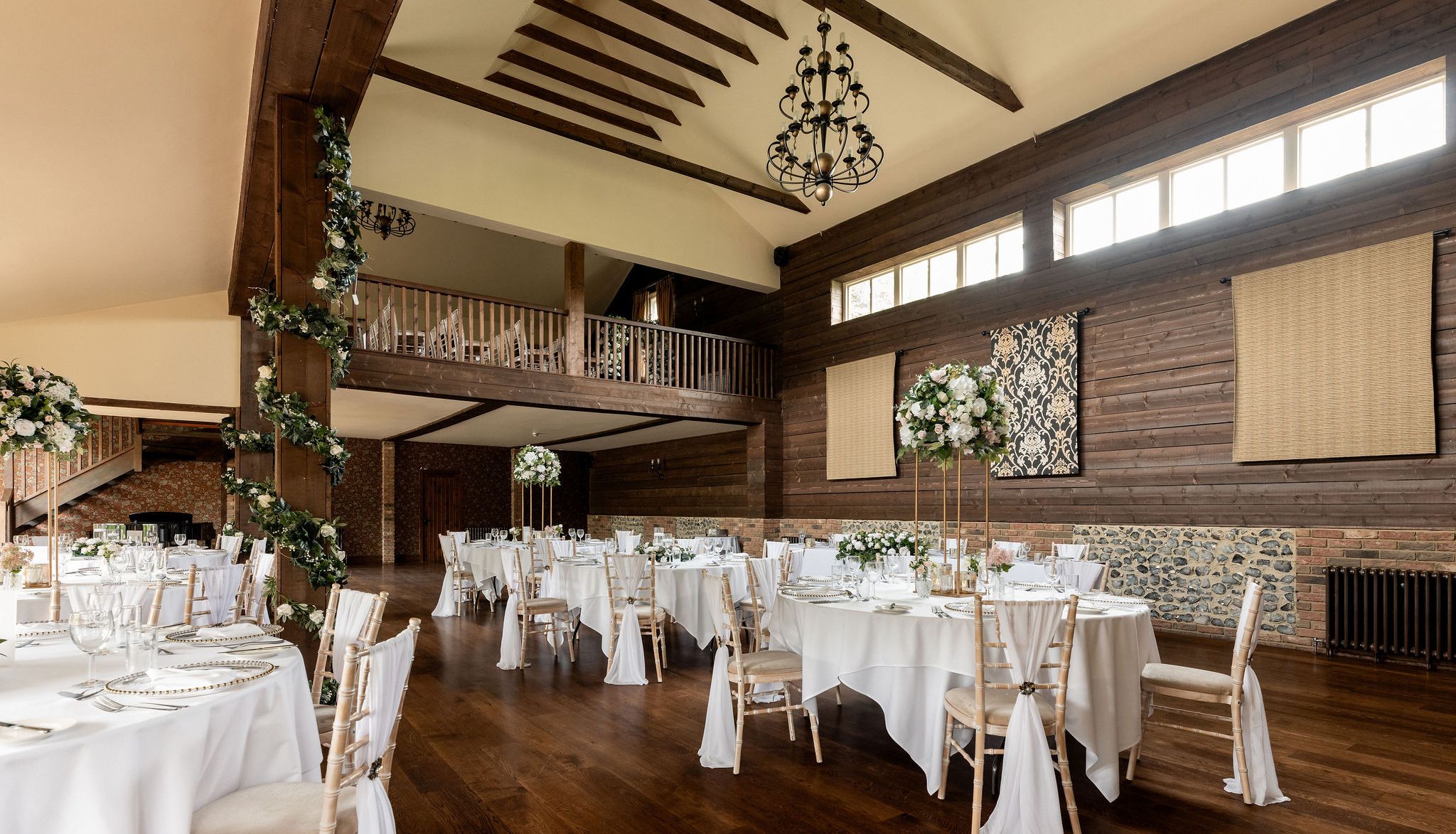 Elegant rustic wedding reception venue with high wooden ceilings, chandeliers, round tables covered in white tablecloths, floral centerpieces, and wooden chairs draped with white fabric.