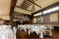 Elegant rustic wedding reception venue with high wooden ceilings, chandeliers, round tables covered in white tablecloths, floral centerpieces, and wooden chairs draped with white fabric.