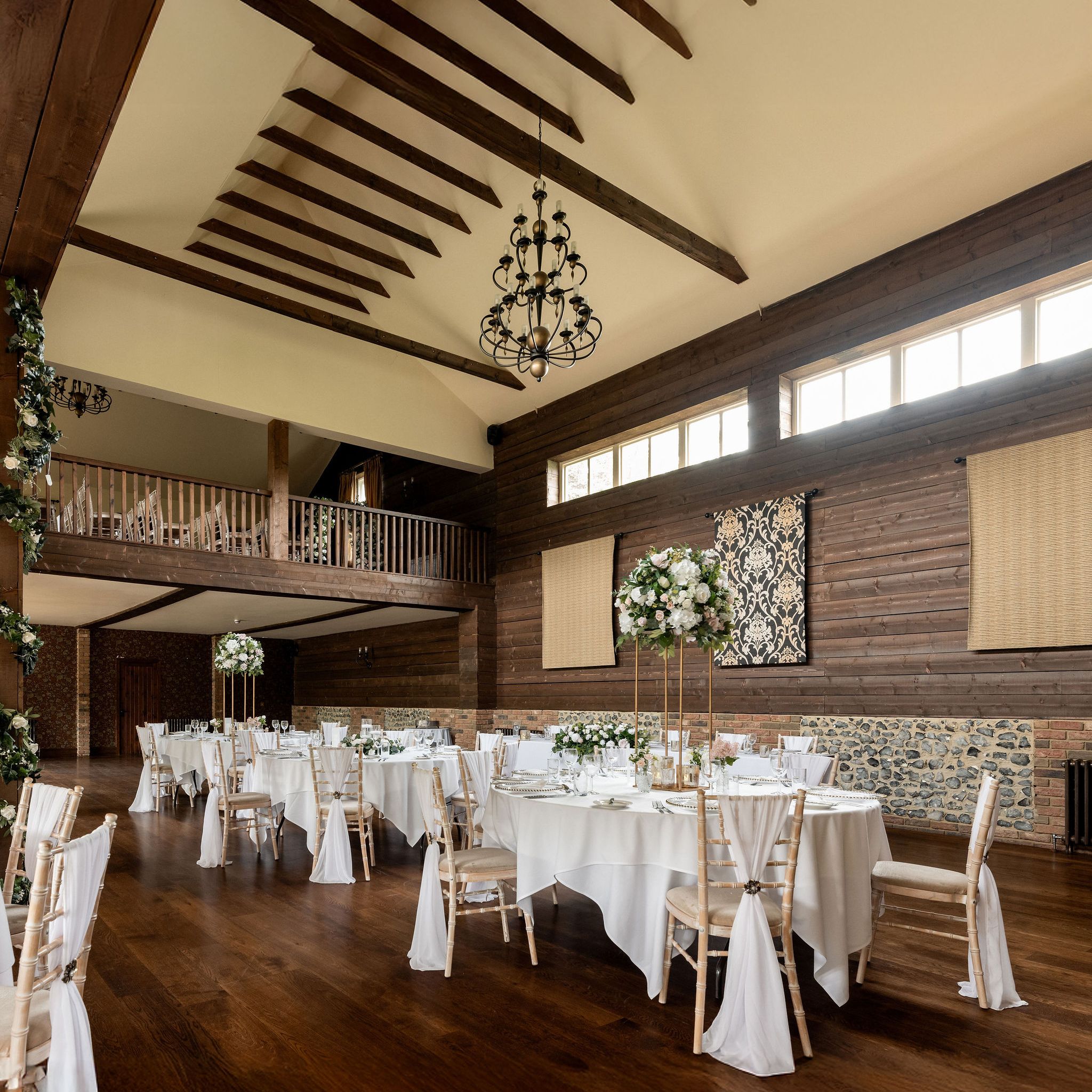Elegant rustic banquet hall with wooden beams, high ceiling, round tables set with white tablecloths, floral centerpieces, and chiavari chairs draped with white fabric.