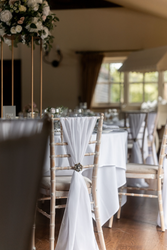 Elegant wedding reception setup with wooden chairs draped in white fabric, decorated with ornate brooches, and tables covered in white linens with floral centerpieces.