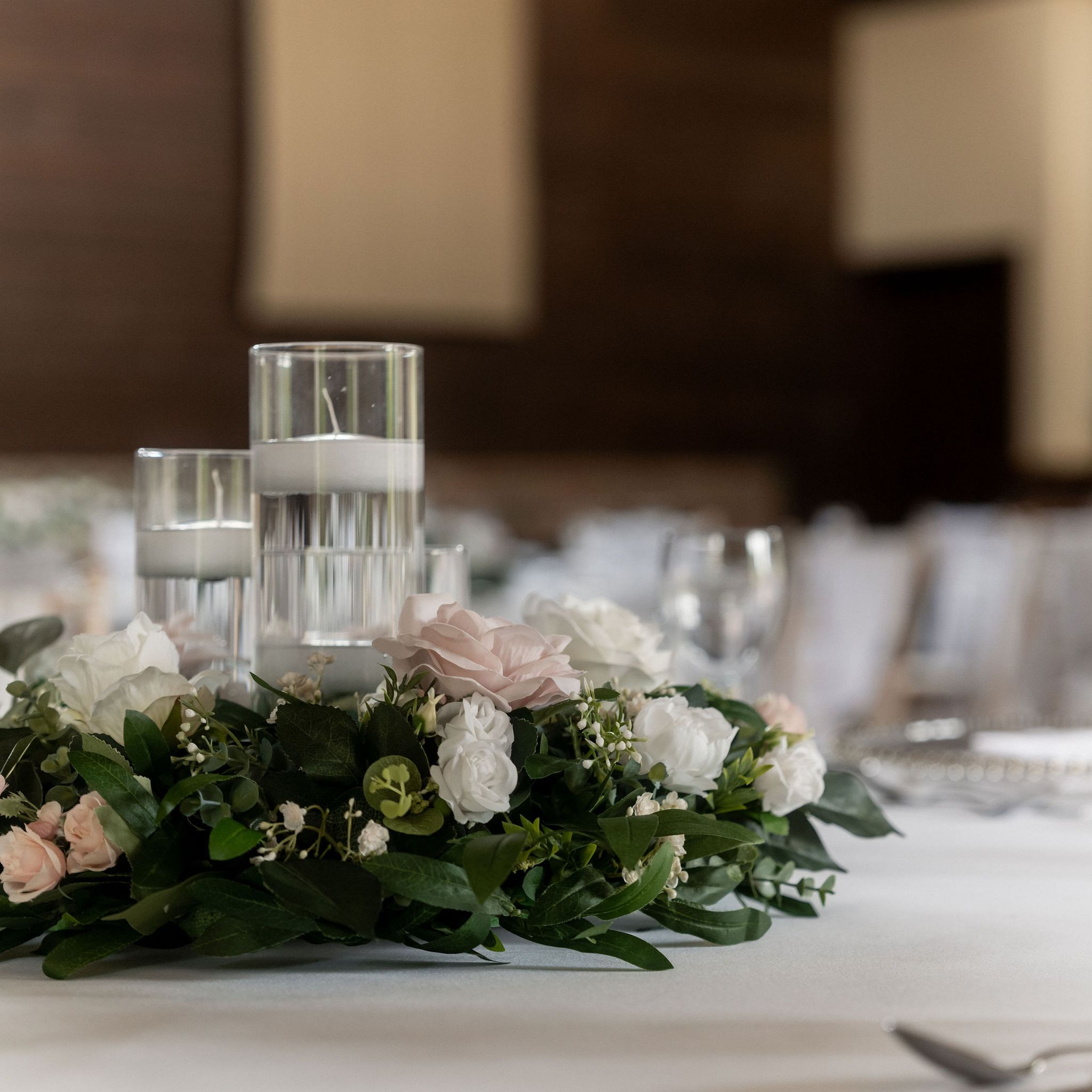 Elegant table centerpiece with floating candles in glass holders surrounded by white and pink flowers and green leaves on a white tablecloth.