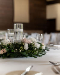Elegant table centerpiece with floating candles in glass holders surrounded by white and pink flowers and green leaves on a white tablecloth.