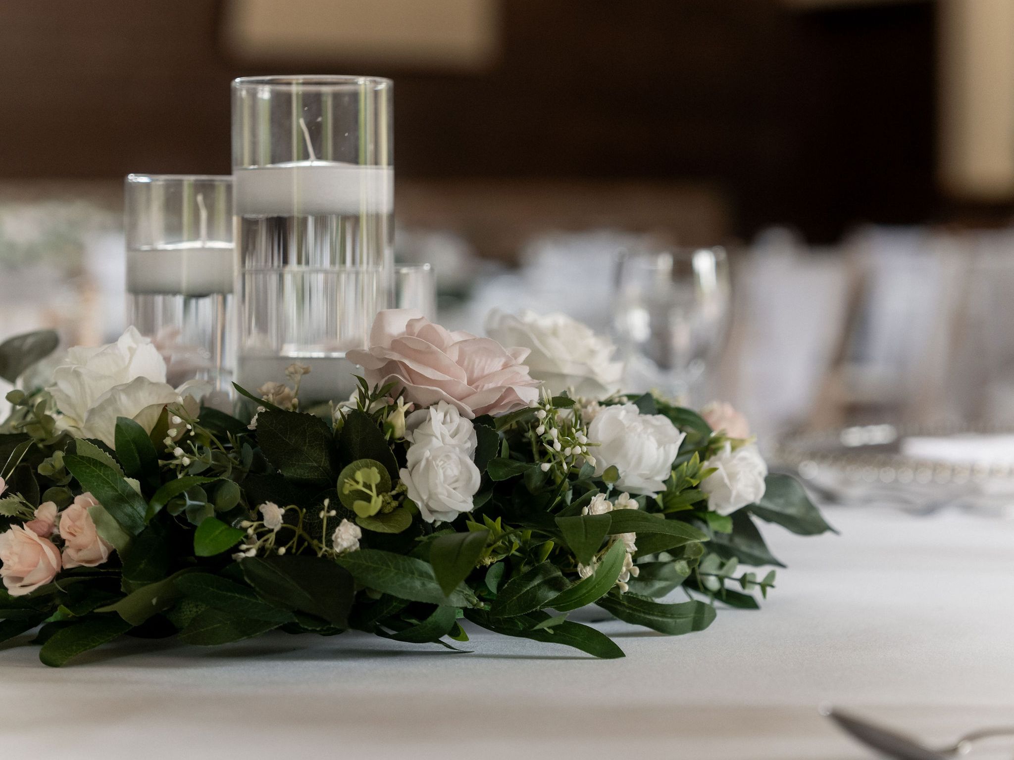 Elegant table centerpiece with floating candles in glass holders surrounded by white and pink flowers and green leaves on a white tablecloth.