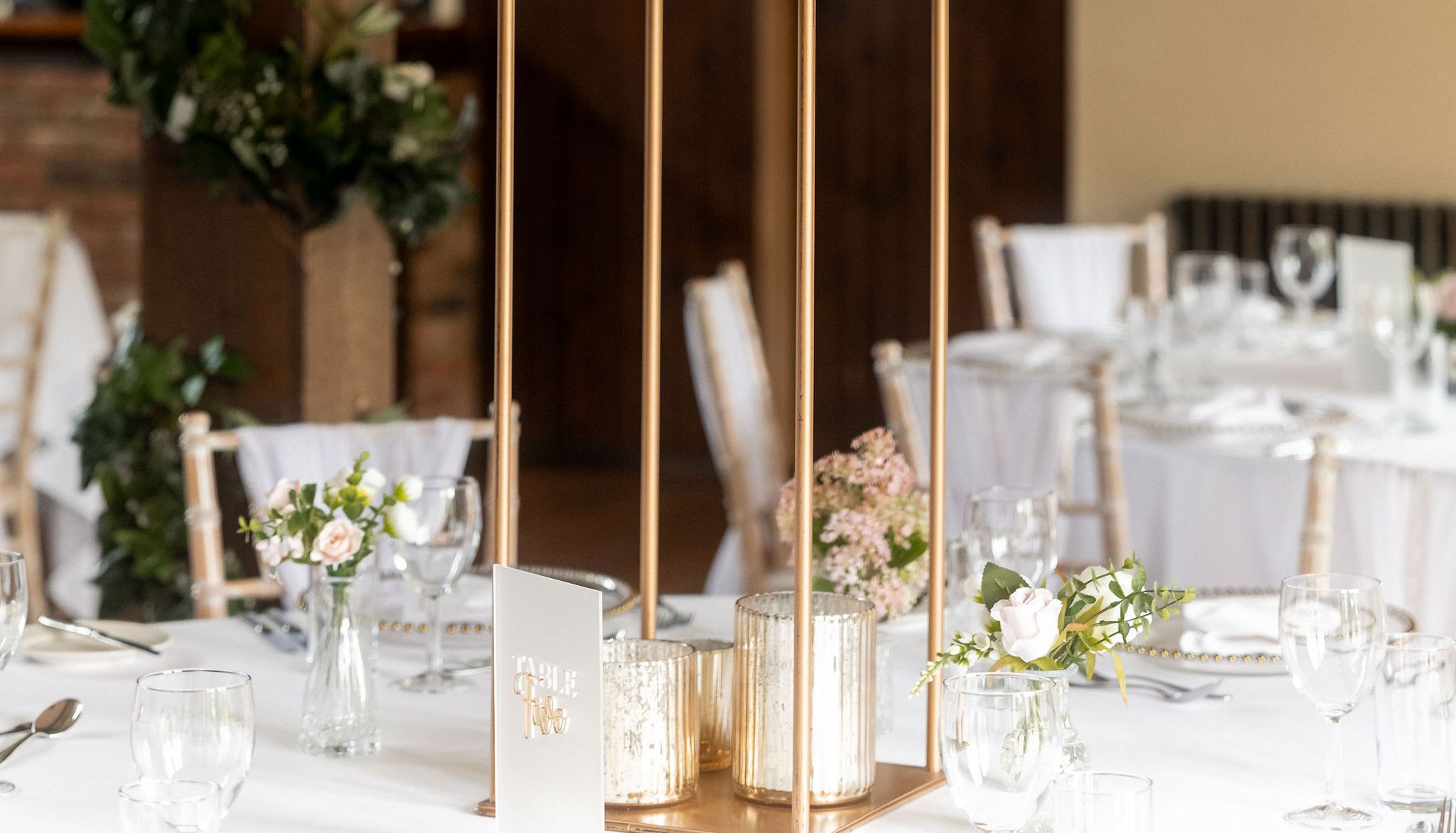 Elegant wedding table centerpiece featuring a tall arrangement of white and blush flowers with greenery on a gold stand, surrounded by glassware and candles in a decorated reception venue.