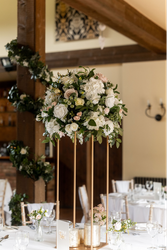 Elegant wedding table centerpiece featuring a tall arrangement of white and blush flowers with greenery on a gold stand, surrounded by glassware and candles in a decorated reception venue.