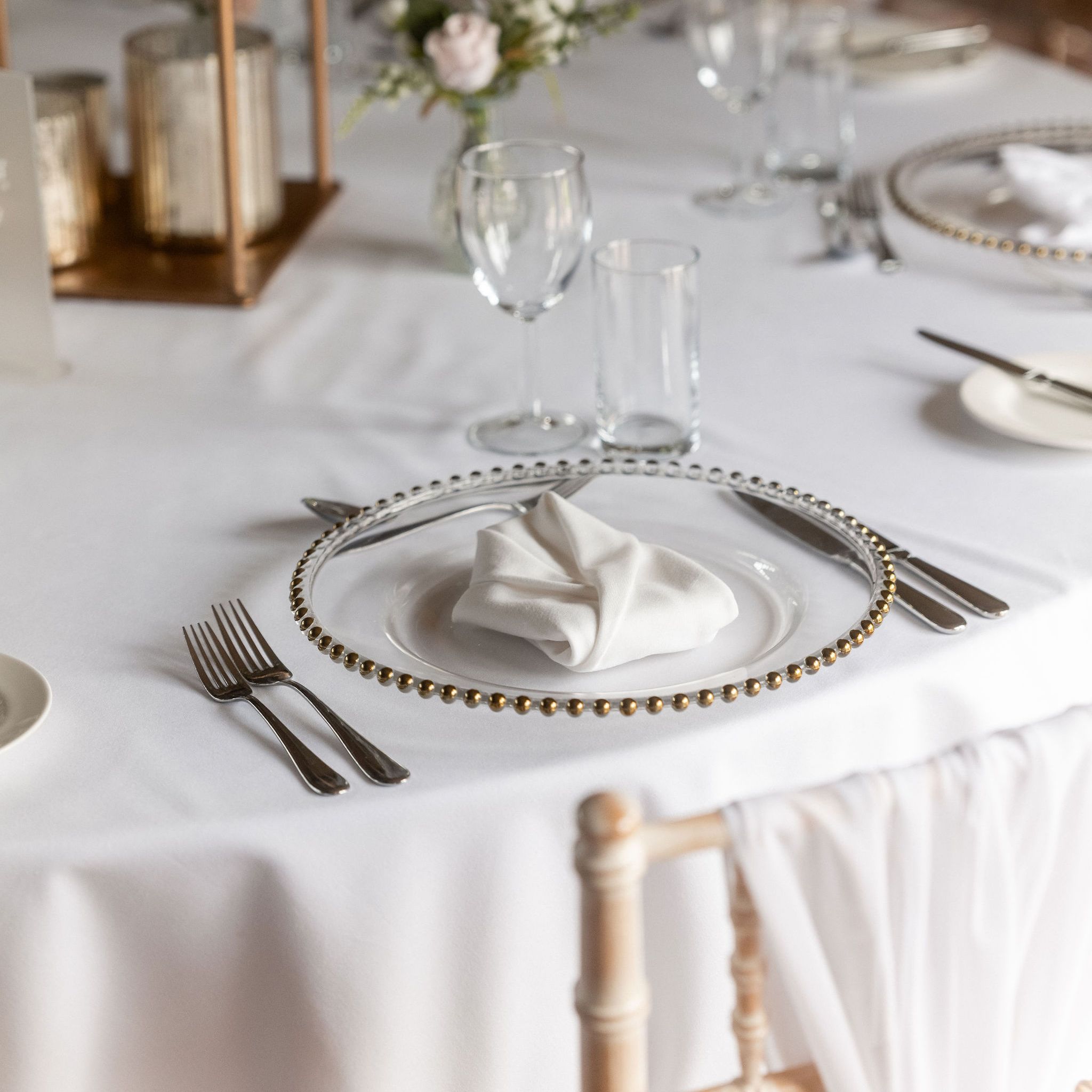 Elegant table setting with a white tablecloth, a decorative plate with a folded white napkin, silverware, glassware, and a small floral centerpiece.