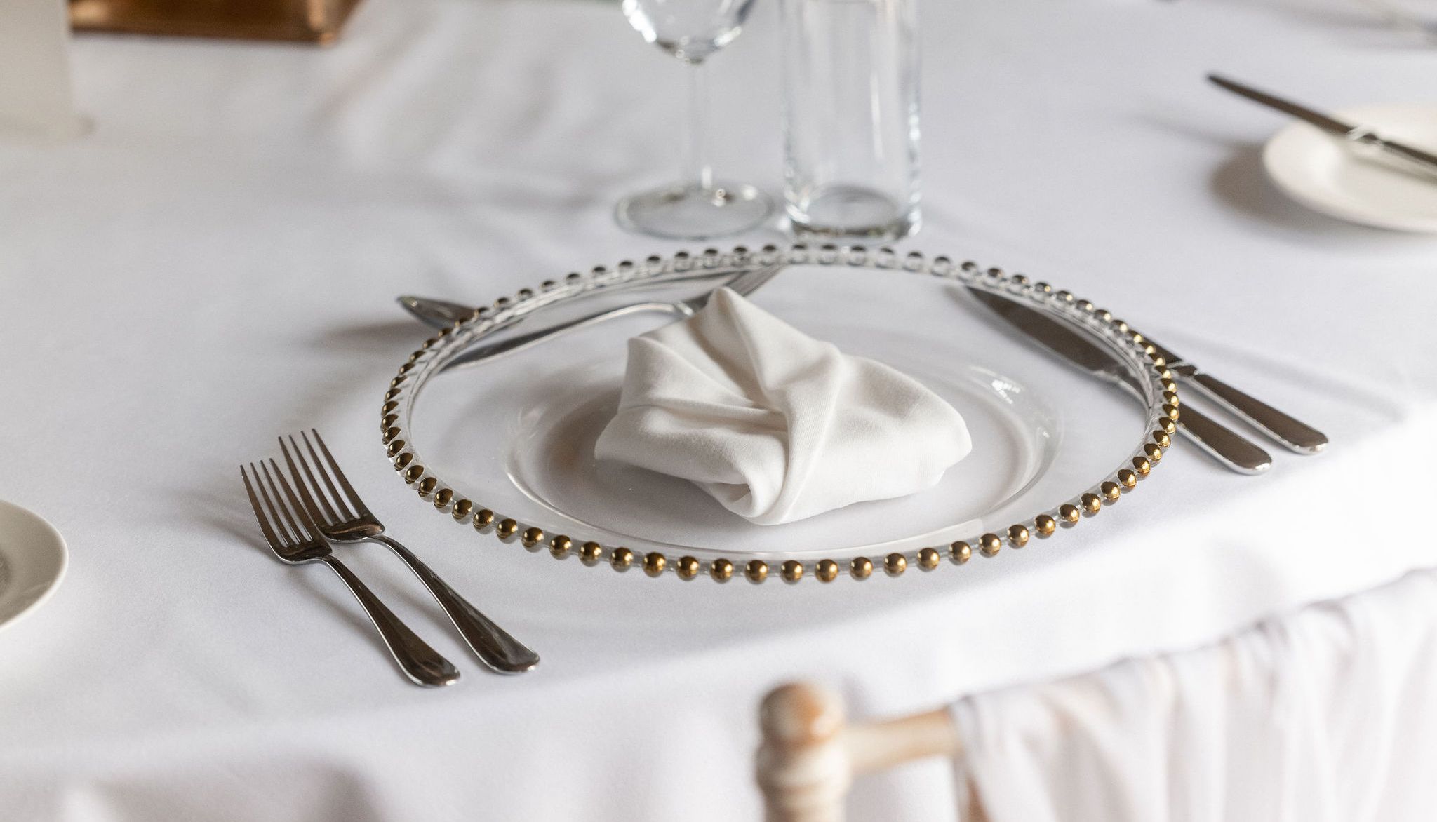 Elegant table setting with a white tablecloth, a decorative plate with a folded white napkin, silverware, glassware, and a small floral centerpiece.
