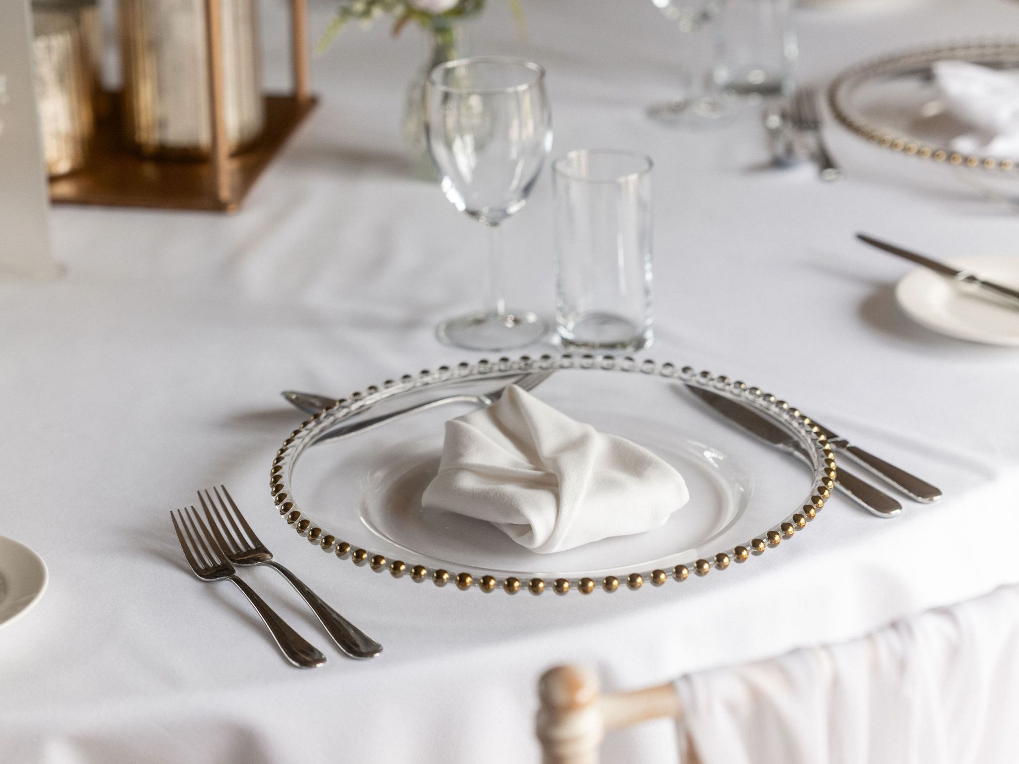 Elegant table setting with a white tablecloth, a decorative plate with a folded white napkin, silverware, glassware, and a small floral centerpiece.